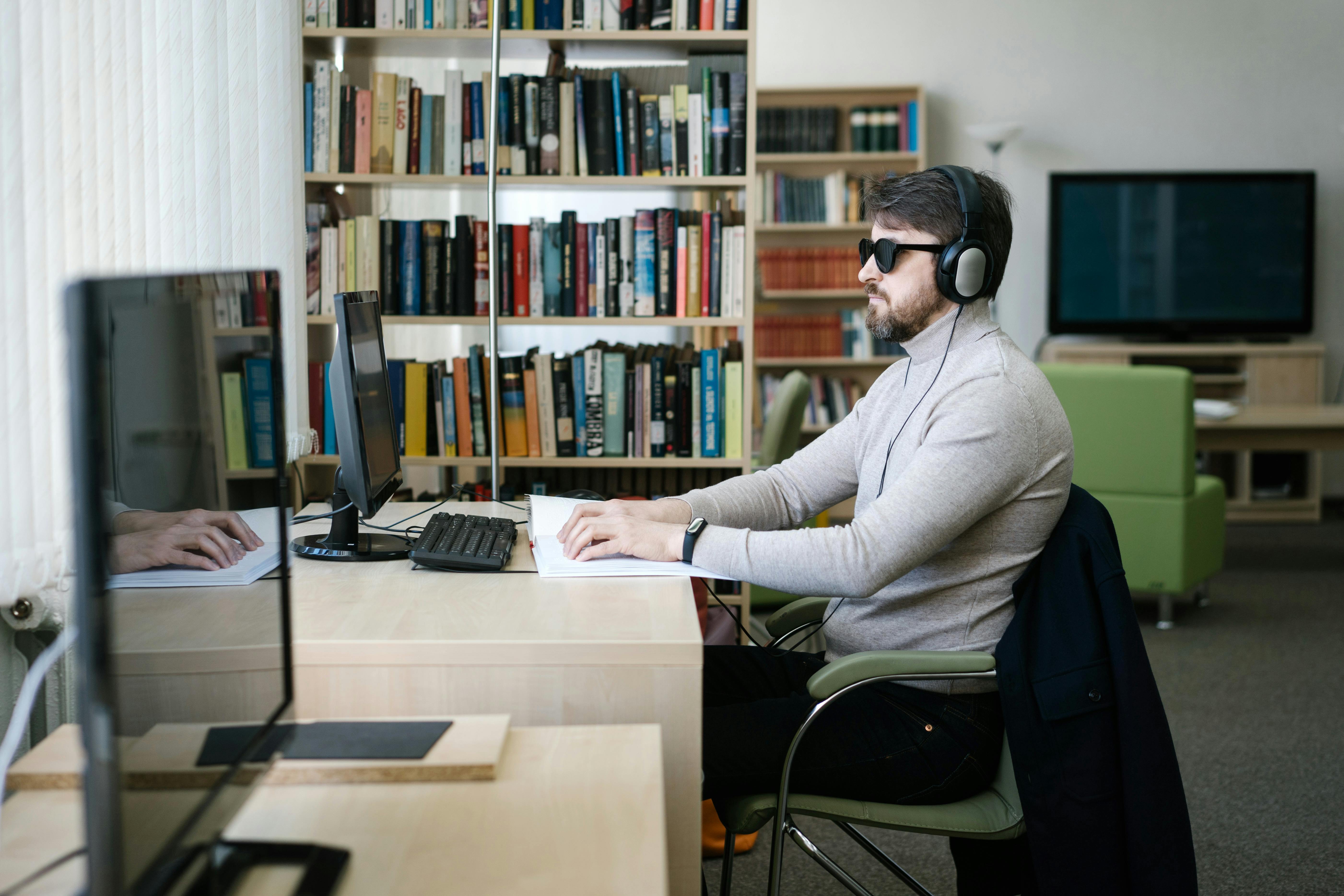 A man wearing sunglasses and headphones reading Braille at a computer