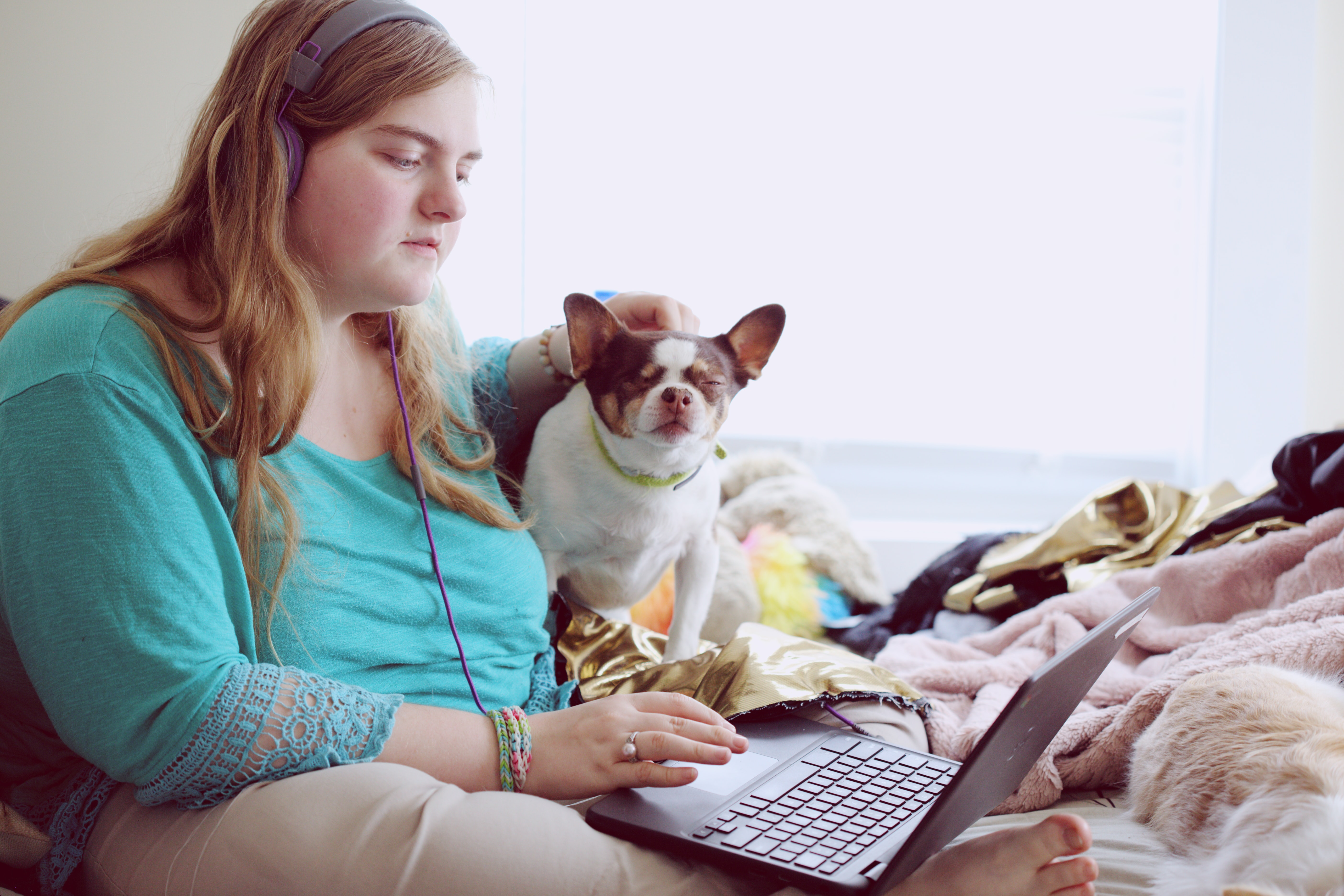 A young woman wearing headphones and using her laptop on her bed while petting her dog.