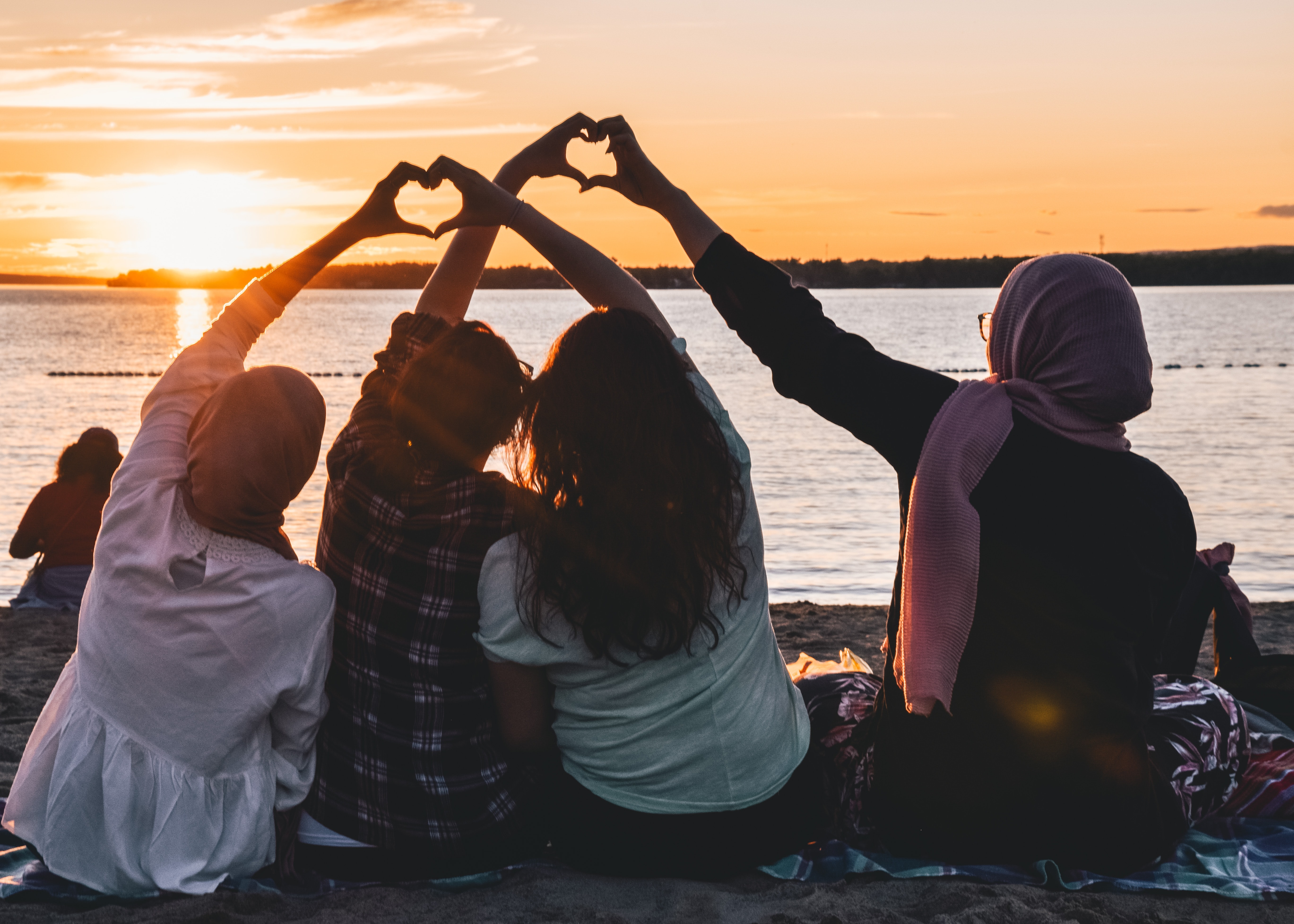 four people sitting on shore forming hearts with their hands during sunset
